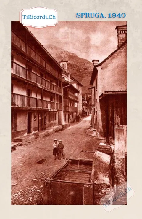 Spruga, 1940: la contrada con le lunghe logge di legno, la fontana in pietra e due bambini in mezzo alla strada